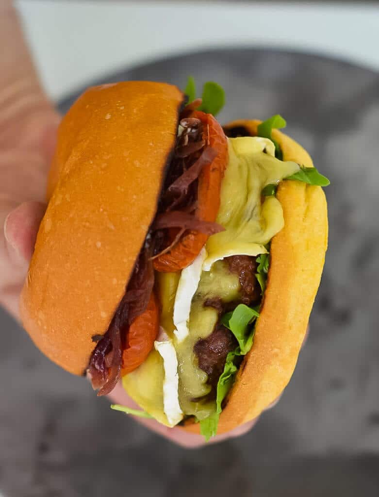 someone holding an organic beef burger with brie, roasted tomatoes & onion marmalade