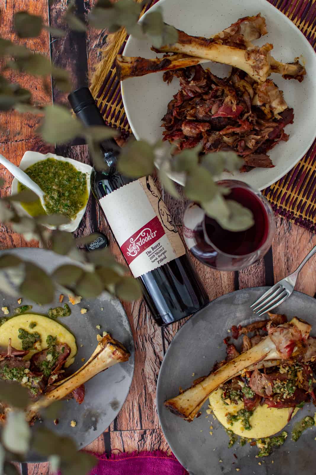 birds eye view of smoked lamb shanks on a plate, wine glass and mollydooker wine 