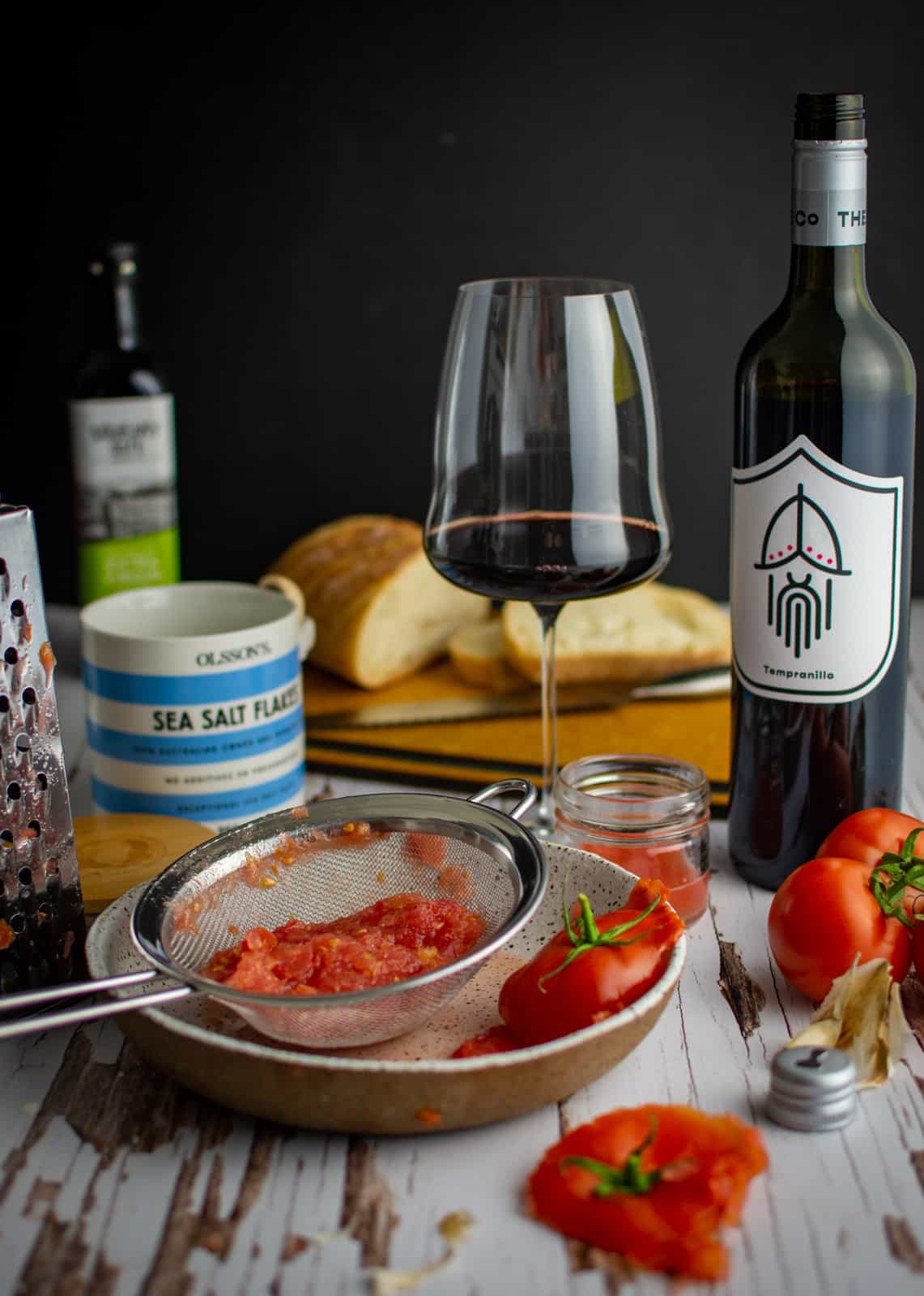 grated tomatoes in a bowl with a sieve.  Red wine, wine glass, salt and bread in background 