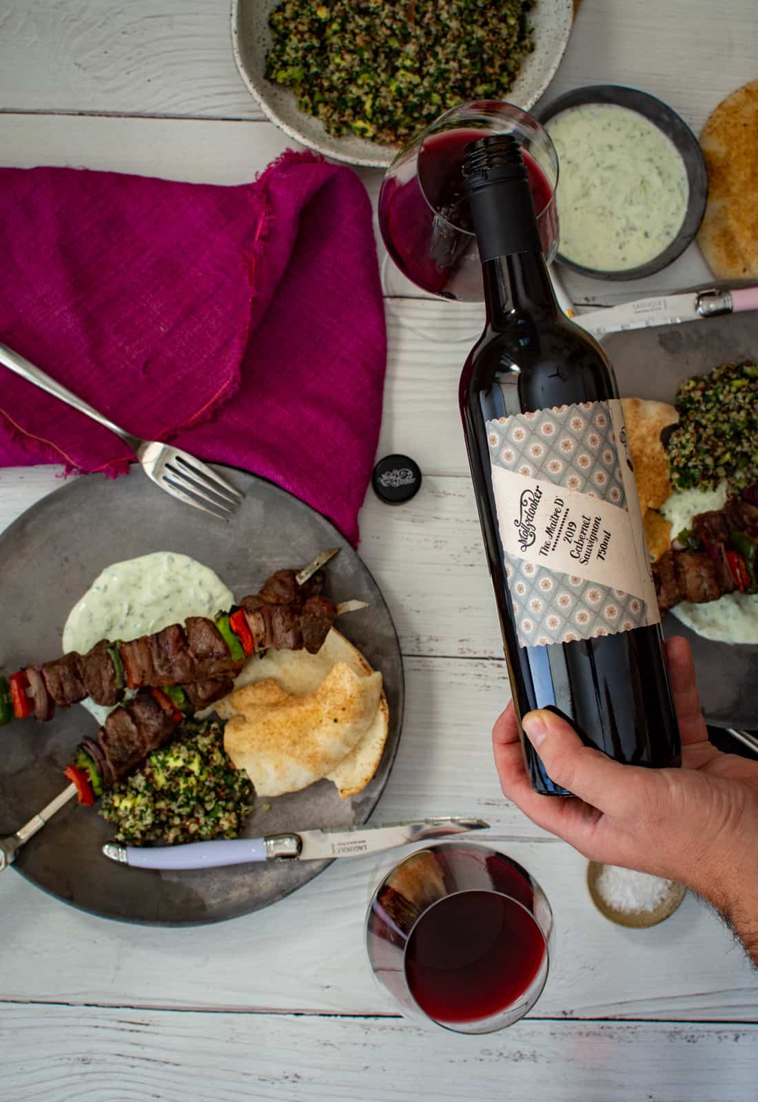 someone pouring Mollydooker red wine with grilled lamb kebabs on plates beside the wine 