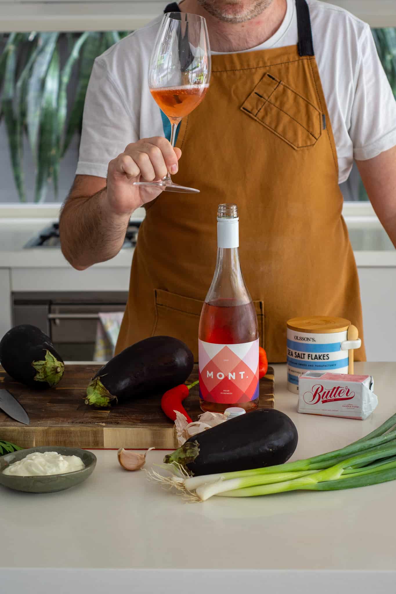 Someone drinking a glass of rose wine in the kitchen with roasted aubergine ingredients on counter top 
