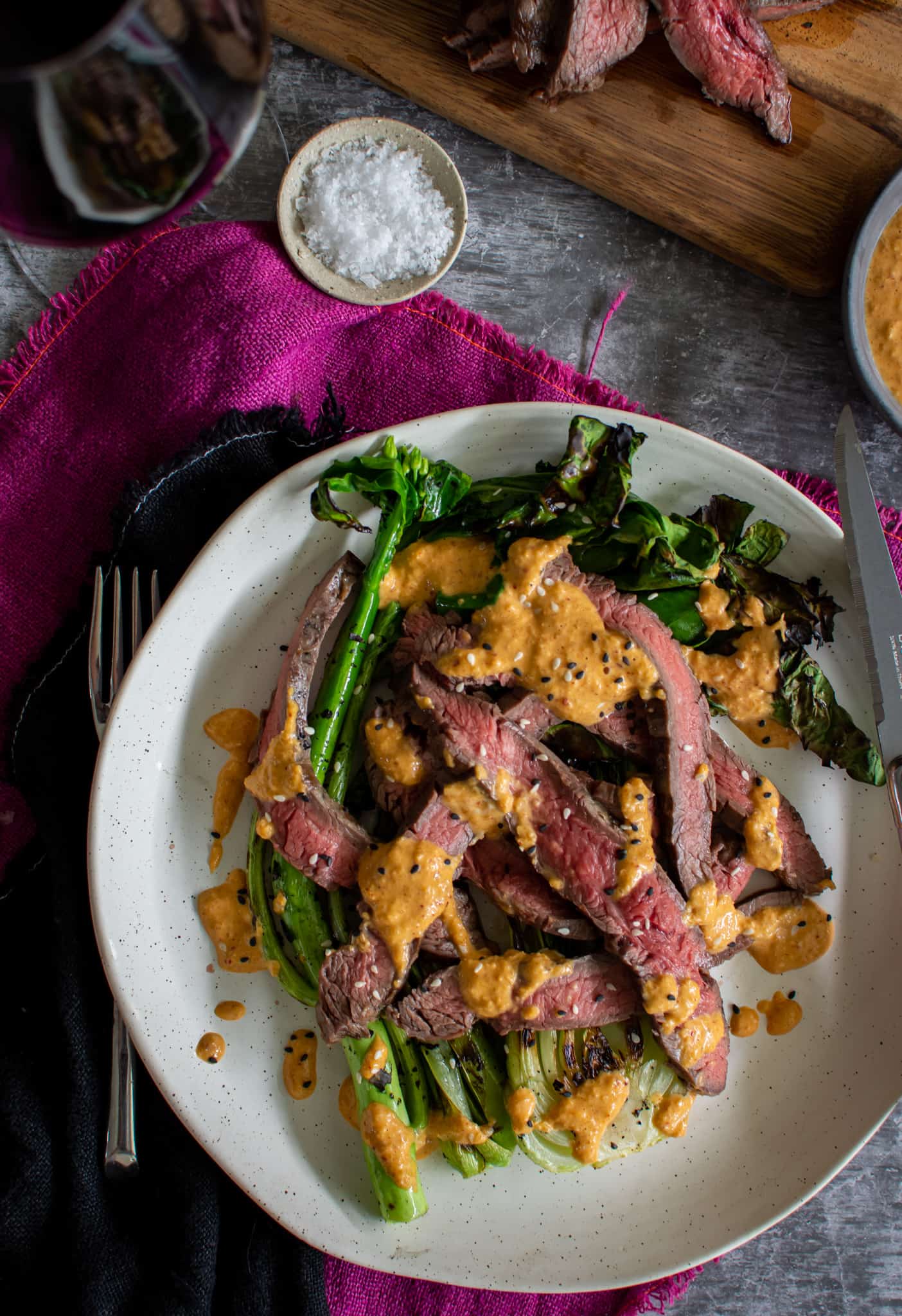 Birdseye view of grilled flank steak, asian greens and kimchi sauce on a white plate with cutlery and napkins beside