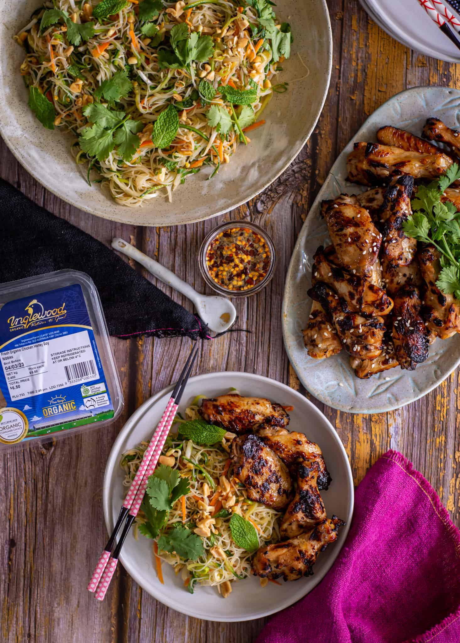 Birdseye view of chicken vermicelli noodle salad in a bowl with pink chopsticks. A plate of grilled chicken nibbles and some napkins are present too 